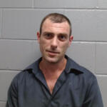 Mugshot-style photo of a man in a dark shirt standing against a gray cinder block wall.