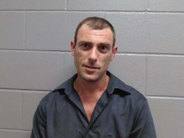 Mugshot-style photo of a man in a dark shirt standing against a gray cinder block wall.