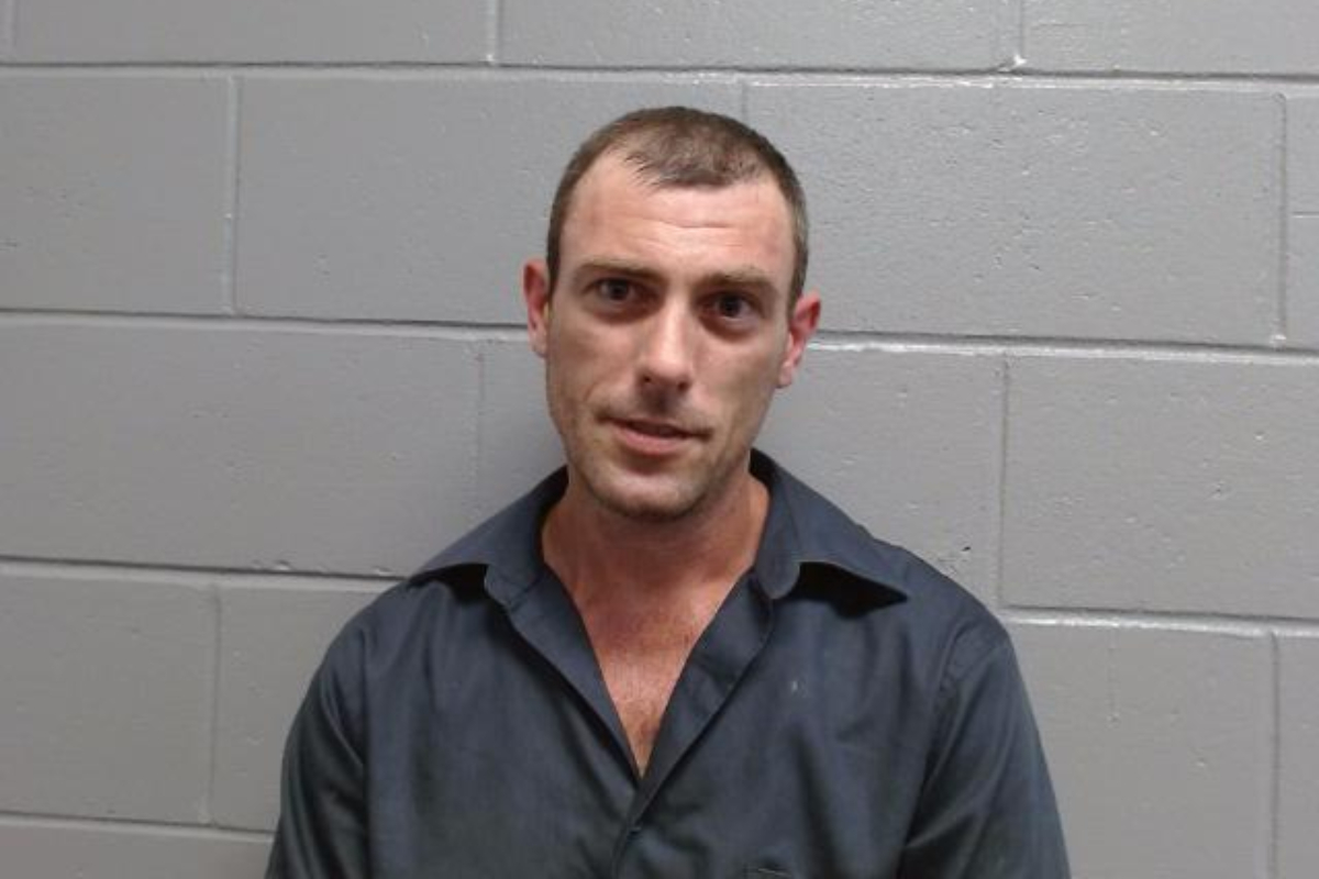 Mugshot-style photo of a man in a dark shirt standing against a gray cinder block wall.