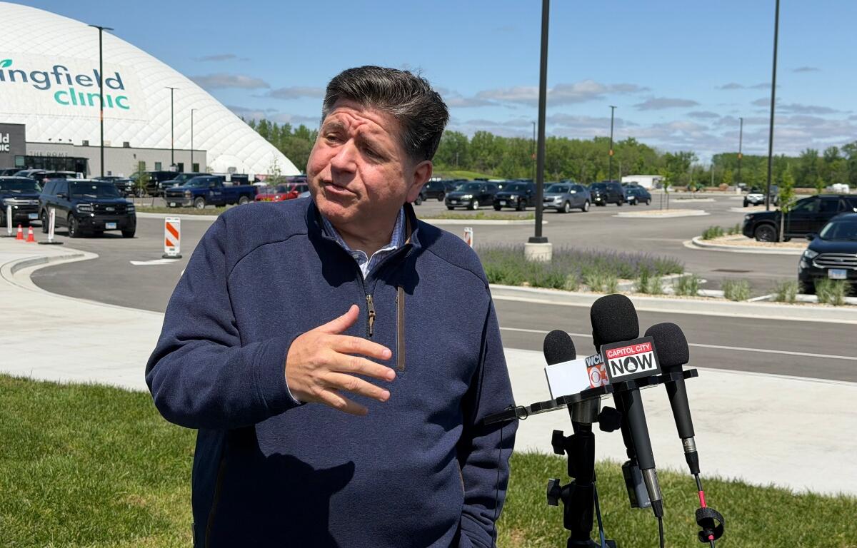 Man in a navy jacket speaks to reporters outside a medical clinic, microphones in front labeled Capitol City Now.