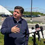 Man in a navy jacket speaks to reporters outside a medical clinic, microphones in front labeled Capitol City Now.