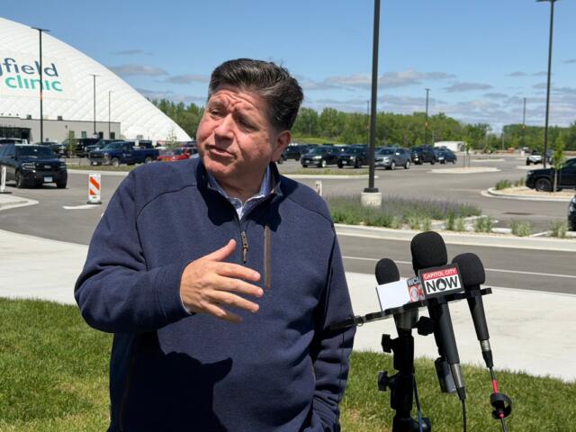 Man in a navy jacket speaks to reporters outside a medical clinic, microphones in front labeled Capitol City Now.
