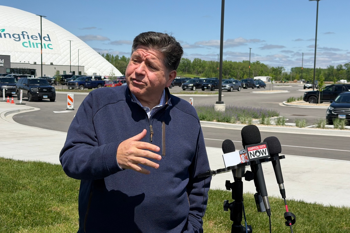 Man in a navy jacket speaks to reporters outside a medical clinic, microphones in front labeled Capitol City Now.