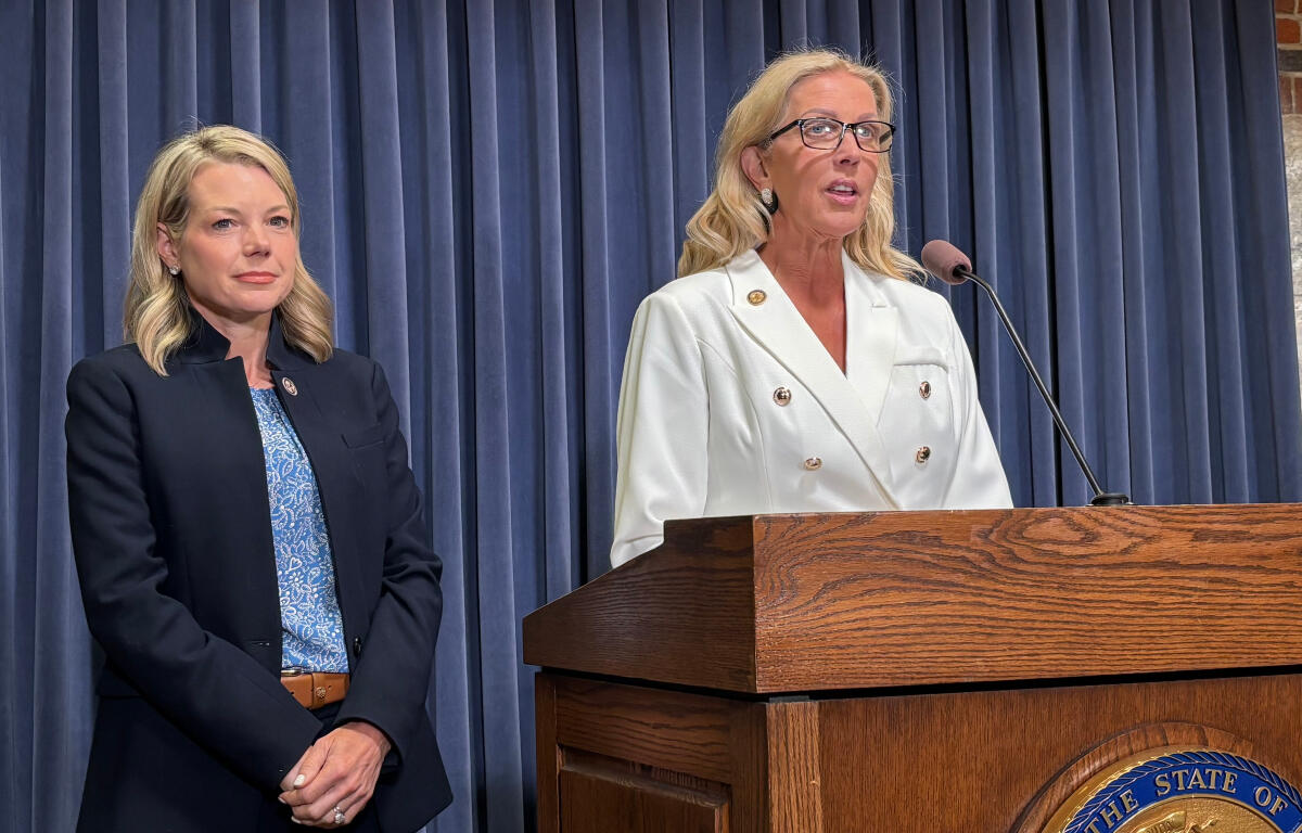 Two women on a stage beside a wooden podium; one speaks into a microphone while wearing a white double-breasted blazer, the other stands in a navy suit behind blue curtains.