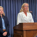 Two women on a stage beside a wooden podium; one speaks into a microphone while wearing a white double-breasted blazer, the other stands in a navy suit behind blue curtains.