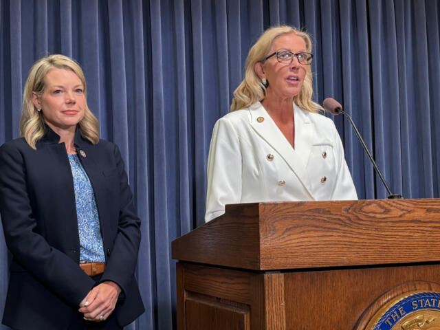 Two women on a stage beside a wooden podium; one speaks into a microphone while wearing a white double-breasted blazer, the other stands in a navy suit behind blue curtains.