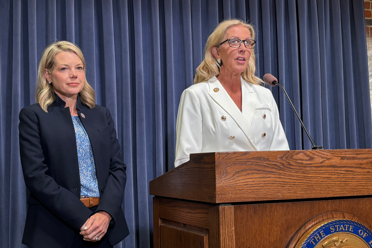 Two women on a stage beside a wooden podium; one speaks into a microphone while wearing a white double-breasted blazer, the other stands in a navy suit behind blue curtains.