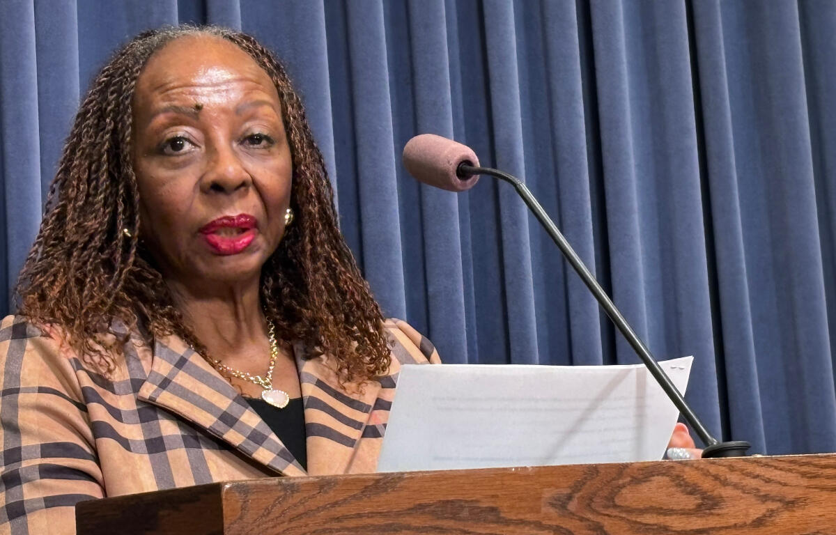 African American woman speaks at a wooden podium with a microphone, holding a document against a blue curtain backdrop.