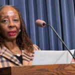 African American woman speaks at a wooden podium with a microphone, holding a document against a blue curtain backdrop.