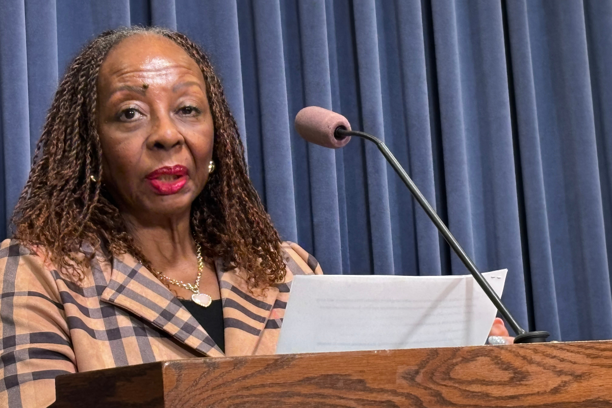 African American woman speaks at a wooden podium with a microphone, holding a document against a blue curtain backdrop.