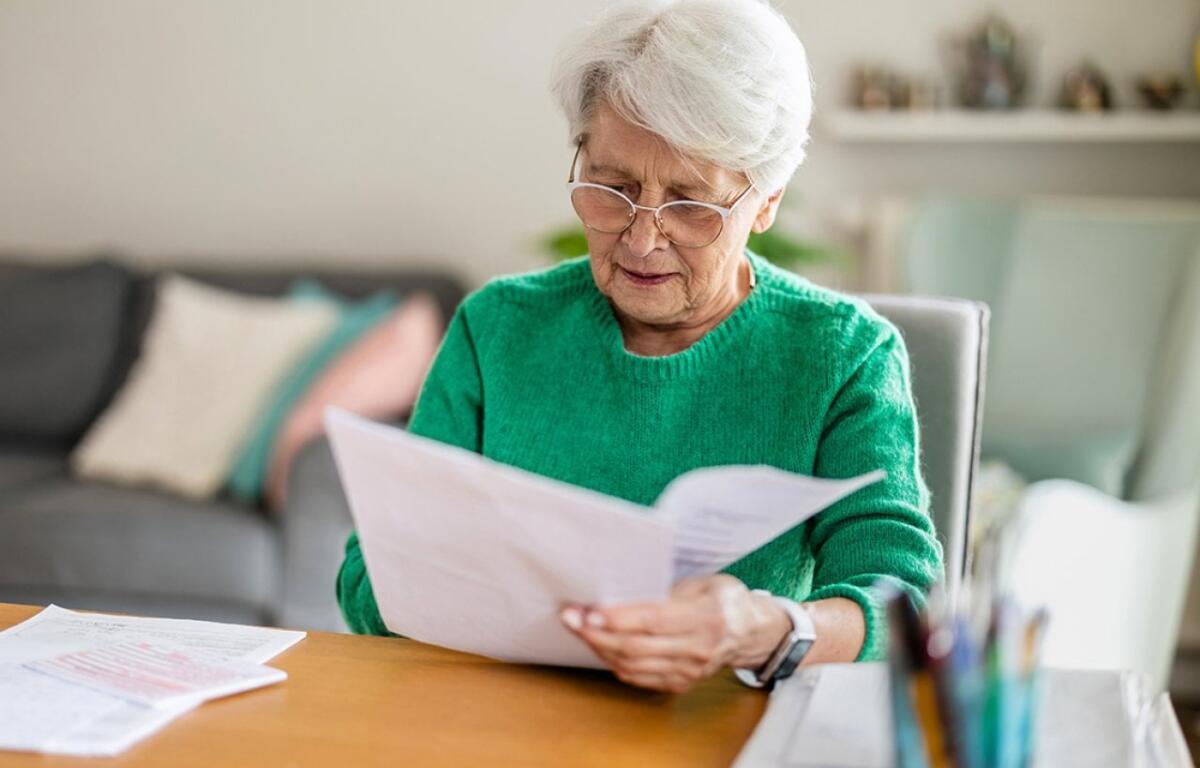 Elderly woman with white hair and glasses reads a stack of papers at a wooden desk in a cozy living room.