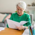 Elderly woman with white hair and glasses reads a stack of papers at a wooden desk in a cozy living room.
