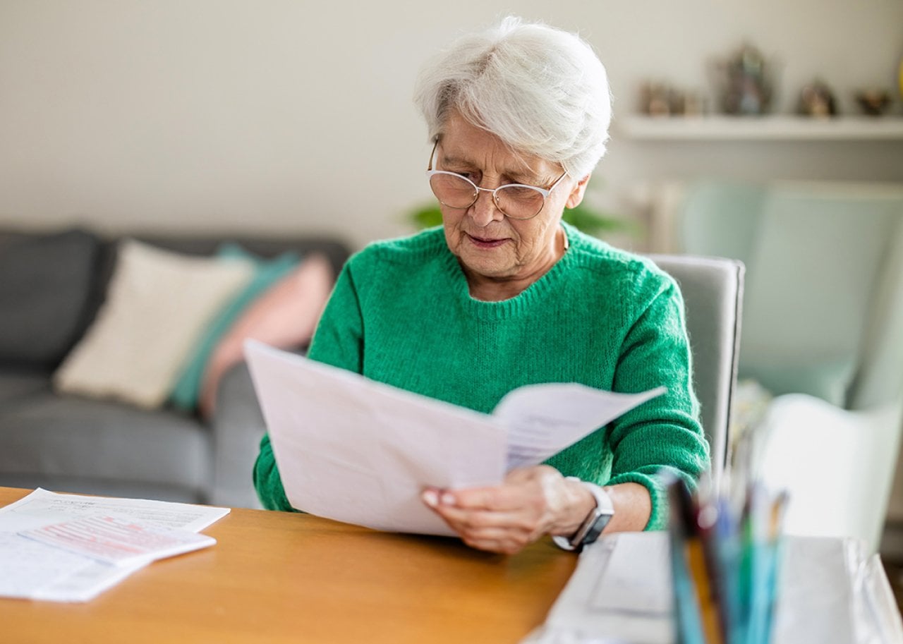 Elderly woman with white hair and glasses reads a stack of papers at a wooden desk in a cozy living room.
