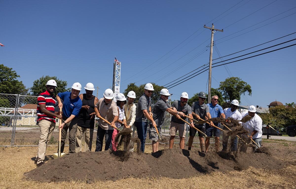Group of people with hard hats using shovels to break ground at a construction site ceremony on a sunny day.