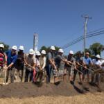 Group of people with hard hats using shovels to break ground at a construction site ceremony on a sunny day.
