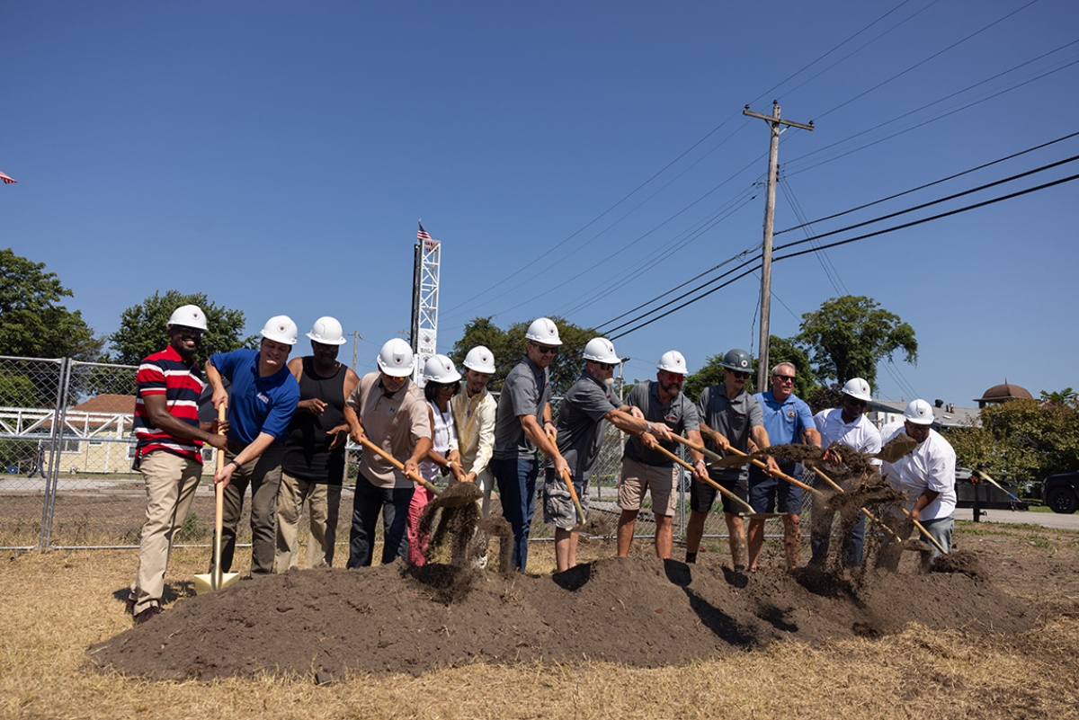 Group of people with hard hats using shovels to break ground at a construction site ceremony on a sunny day.