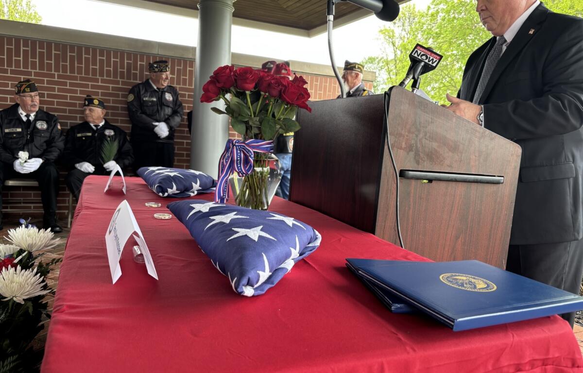 Male speaker in a dark suit at podium with microphones during a veterans' ceremony, floral arrangements nearby.