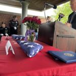 Male speaker in a dark suit at podium with microphones during a veterans' ceremony, floral arrangements nearby.
