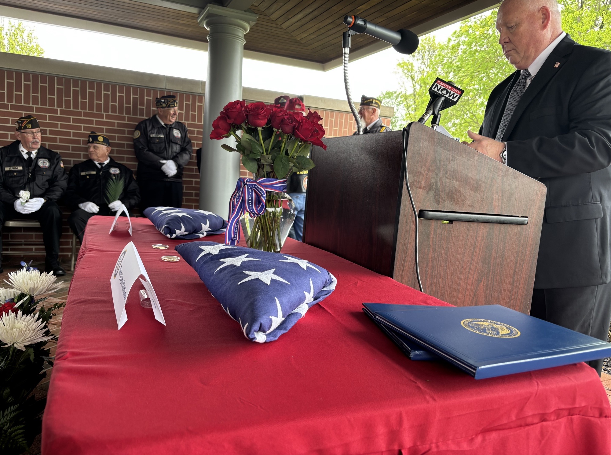 Male speaker in a dark suit at podium with microphones during a veterans' ceremony, floral arrangements nearby.