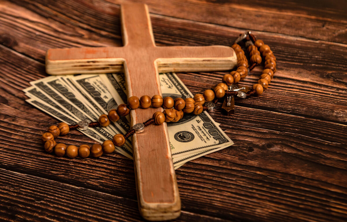 Wooden cross with a bead rosary draped over scattered $100 bills on a rustic wooden surface, suggesting faith and money.
