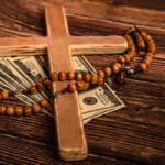 Wooden cross with a bead rosary draped over scattered $100 bills on a rustic wooden surface, suggesting faith and money.