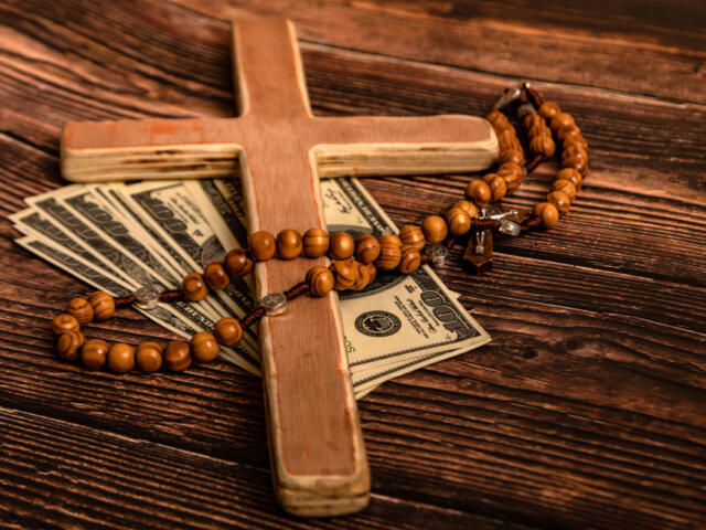 Wooden cross with a bead rosary draped over scattered $100 bills on a rustic wooden surface, suggesting faith and money.