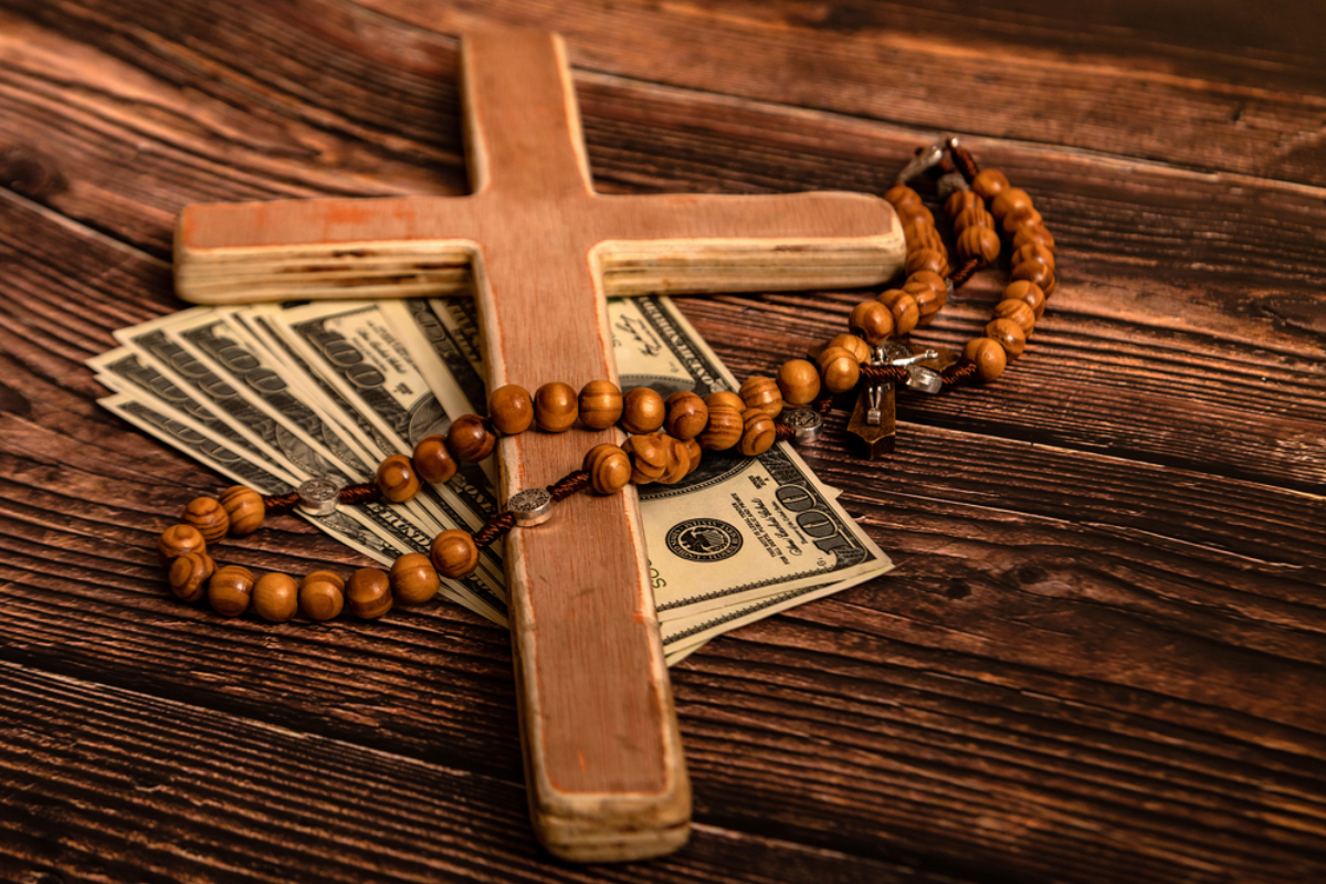 Wooden cross with a bead rosary draped over scattered $100 bills on a rustic wooden surface, suggesting faith and money.
