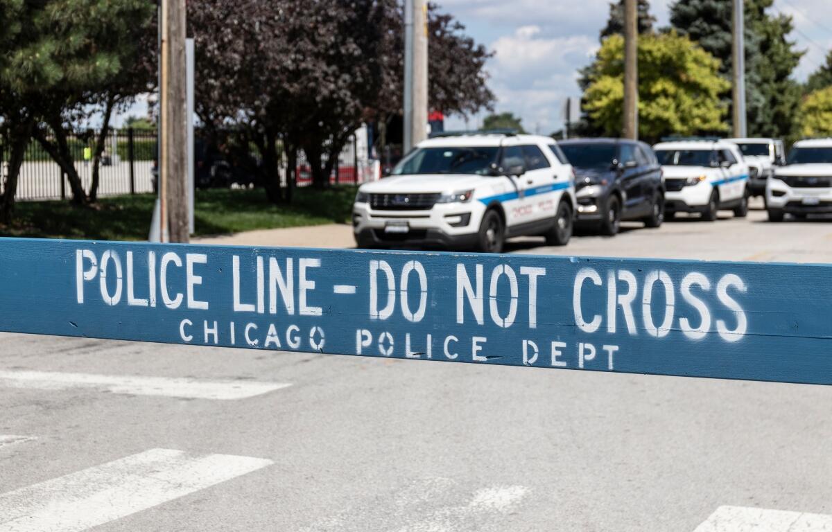 Blue police barrier with white stencil text: 'POLICE LINE - DO NOT CROSS' and 'CHICAGO POLICE DEPT'; police cars in background indicating a crime scene or checkpoint.
