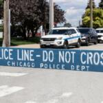 Blue police barrier with white stencil text: 'POLICE LINE - DO NOT CROSS' and 'CHICAGO POLICE DEPT'; police cars in background indicating a crime scene or checkpoint.