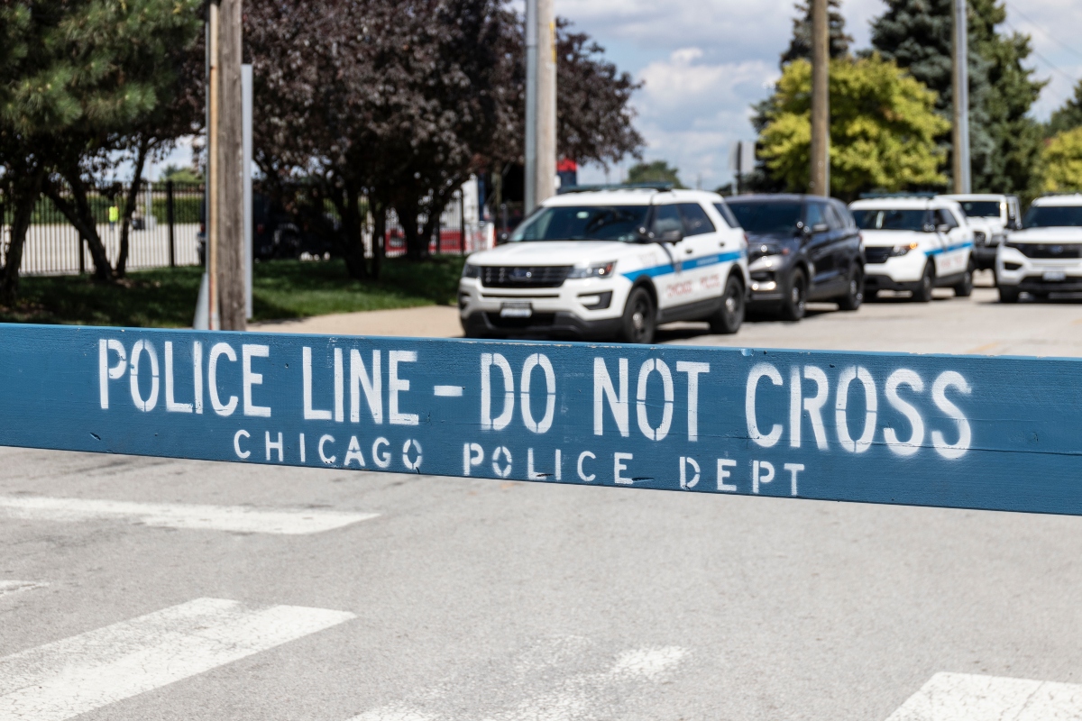 Blue police barrier with white stencil text: 'POLICE LINE - DO NOT CROSS' and 'CHICAGO POLICE DEPT'; police cars in background indicating a crime scene or checkpoint.
