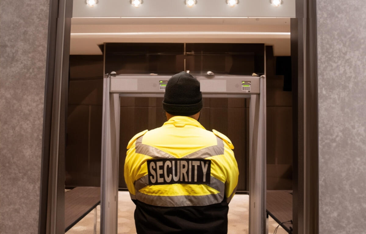 Security guard in a neon-yellow jacket standing at a metal detector, viewed from behind.