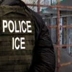 An Immigration and Customs Enforcement (ICE) officer stands in front of a dilapidated detention facility gate.