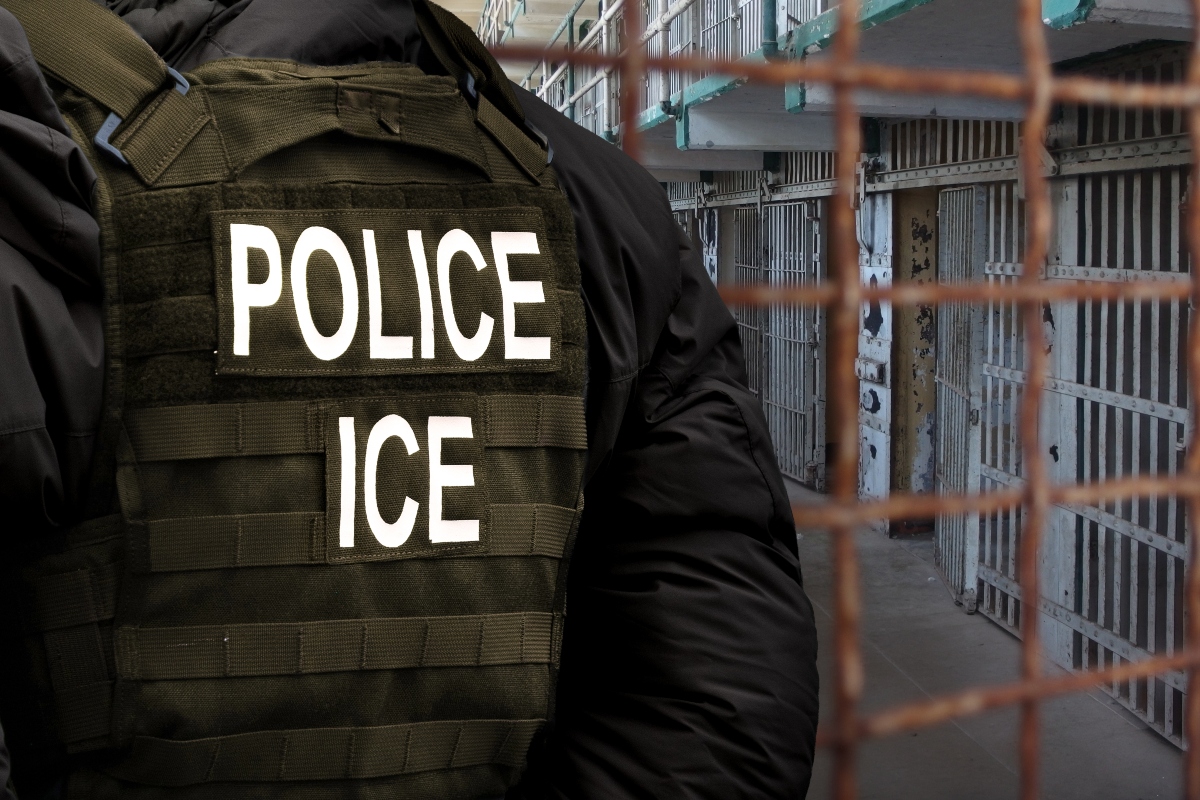 An Immigration and Customs Enforcement (ICE) officer stands in front of a dilapidated detention facility gate.