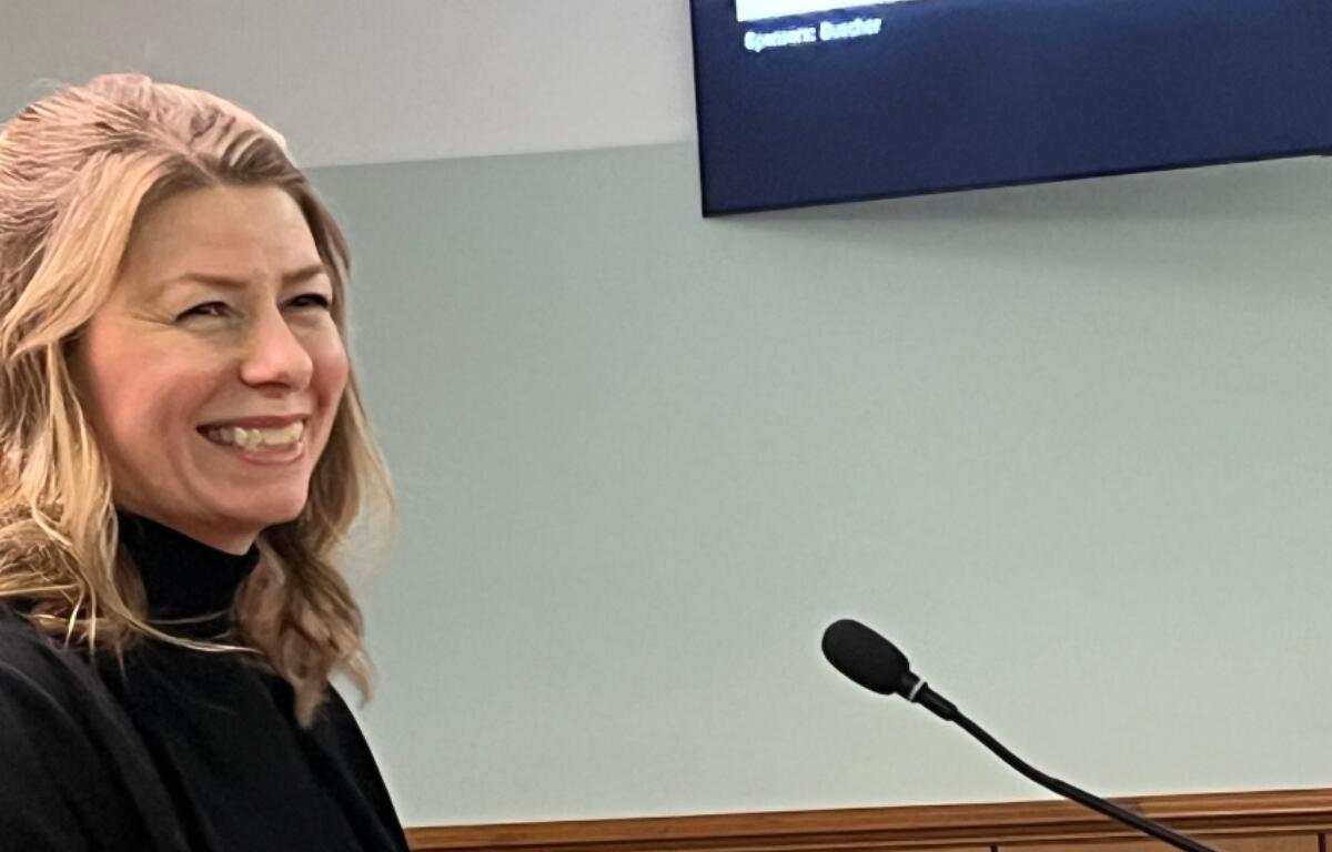 Smiling blonde woman in a black top speaking at a podium with a microphone in a meeting room.