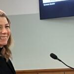 Smiling blonde woman in a black top speaking at a podium with a microphone in a meeting room.
