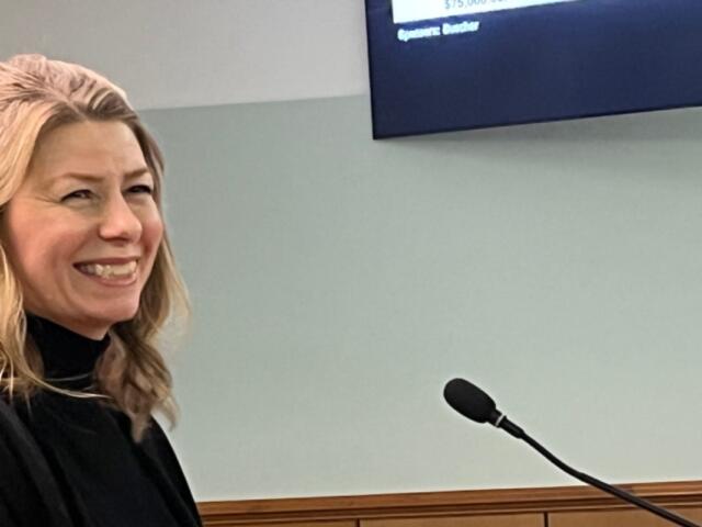 Smiling blonde woman in a black top speaking at a podium with a microphone in a meeting room.