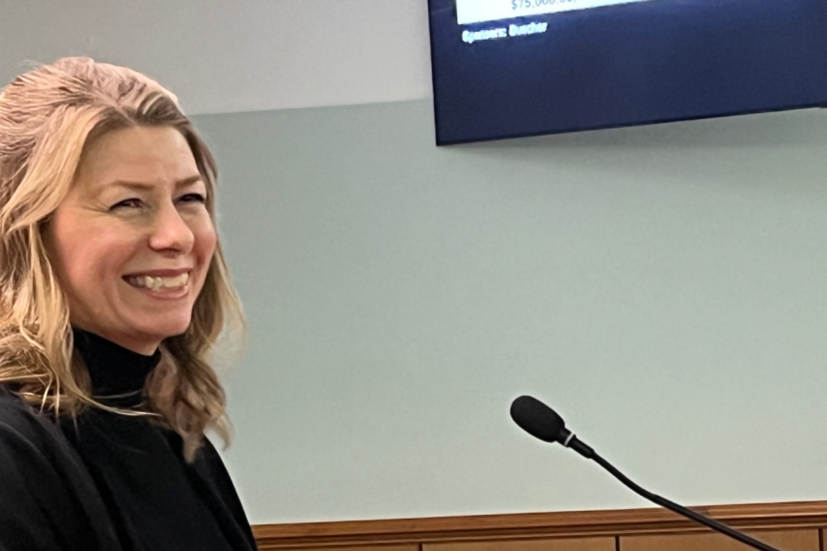 Smiling blonde woman in a black top speaking at a podium with a microphone in a meeting room.