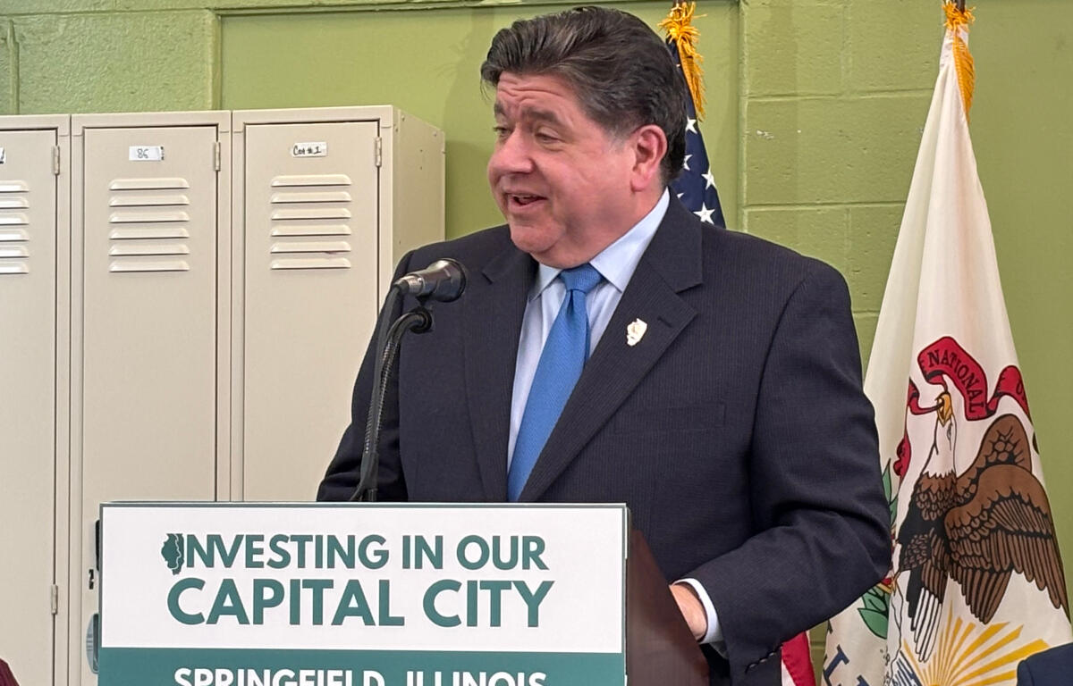 Man in a dark suit and blue tie speaks at a podium with a sign reading 'Investing in Our Capital City' in Springfield, Illinois, flanked by flags and lockers in the background.