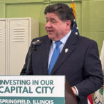 Man in a dark suit and blue tie speaks at a podium with a sign reading 'Investing in Our Capital City' in Springfield, Illinois, flanked by flags and lockers in the background.