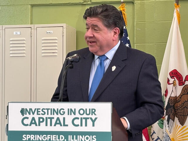Man in a dark suit and blue tie speaks at a podium with a sign reading 'Investing in Our Capital City' in Springfield, Illinois, flanked by flags and lockers in the background.