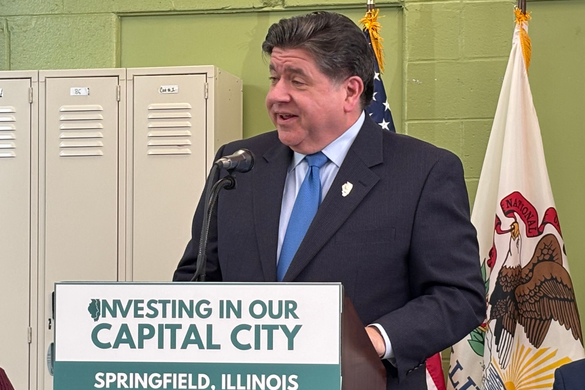Man in a dark suit and blue tie speaks at a podium with a sign reading 'Investing in Our Capital City' in Springfield, Illinois, flanked by flags and lockers in the background.