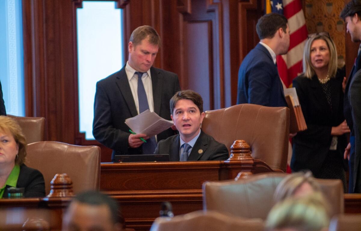 Man in a suit sits at a wooden desk in a formal government chamber, while colleagues chat in the background near an American flag.