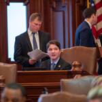 Man in a suit sits at a wooden desk in a formal government chamber, while colleagues chat in the background near an American flag.