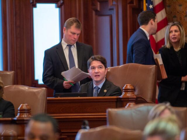 Man in a suit sits at a wooden desk in a formal government chamber, while colleagues chat in the background near an American flag.