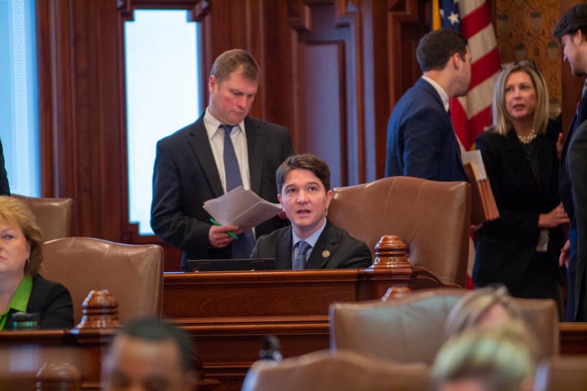 Man in a suit sits at a wooden desk in a formal government chamber, while colleagues chat in the background near an American flag.