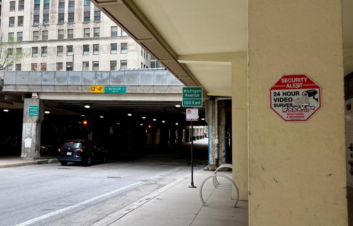 Urban street scene beneath an elevated highway, with a dark car parked near concrete pillars and green Michigan Avenue signs nearby; a red Security Alert sign is mounted on a beige column.