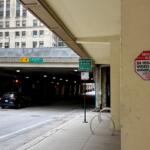Urban street scene beneath an elevated highway, with a dark car parked near concrete pillars and green Michigan Avenue signs nearby; a red Security Alert sign is mounted on a beige column.