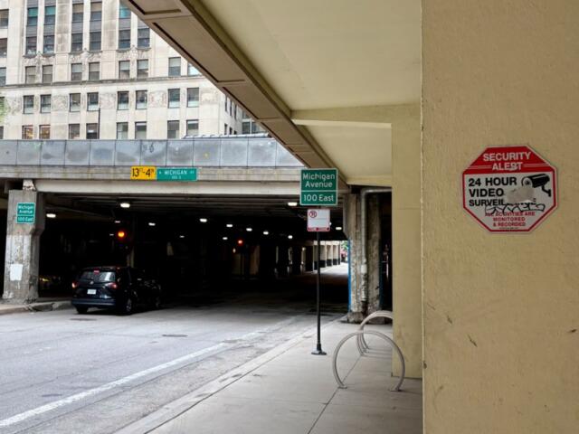 Urban street scene beneath an elevated highway, with a dark car parked near concrete pillars and green Michigan Avenue signs nearby; a red Security Alert sign is mounted on a beige column.