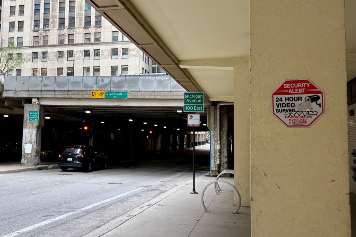 Urban street scene beneath an elevated highway, with a dark car parked near concrete pillars and green Michigan Avenue signs nearby; a red Security Alert sign is mounted on a beige column.
