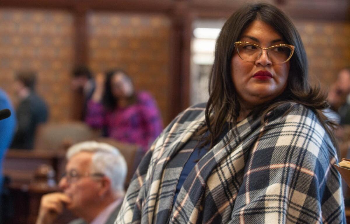 Woman with dark hair and glasses wearing a blue plaid shawl, looking to the right at a meeting.